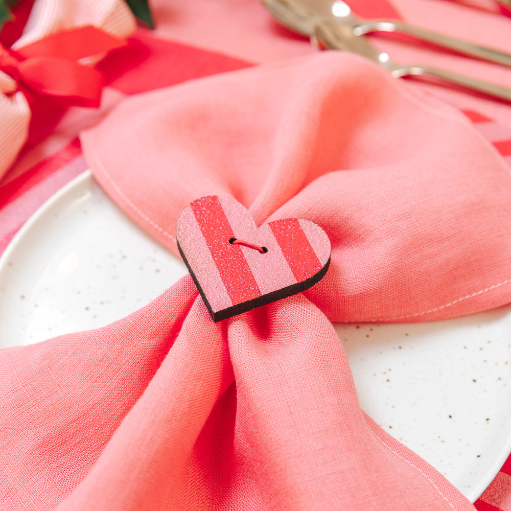 Pink napkin with a heart-shaped cork clip on a white plate, surrounded by red ribbons and gold cutlery.