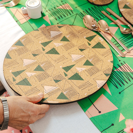 Person holding a geometric-patterned cork placemat over a green tablecloth with a geometric midcentury inspired design.