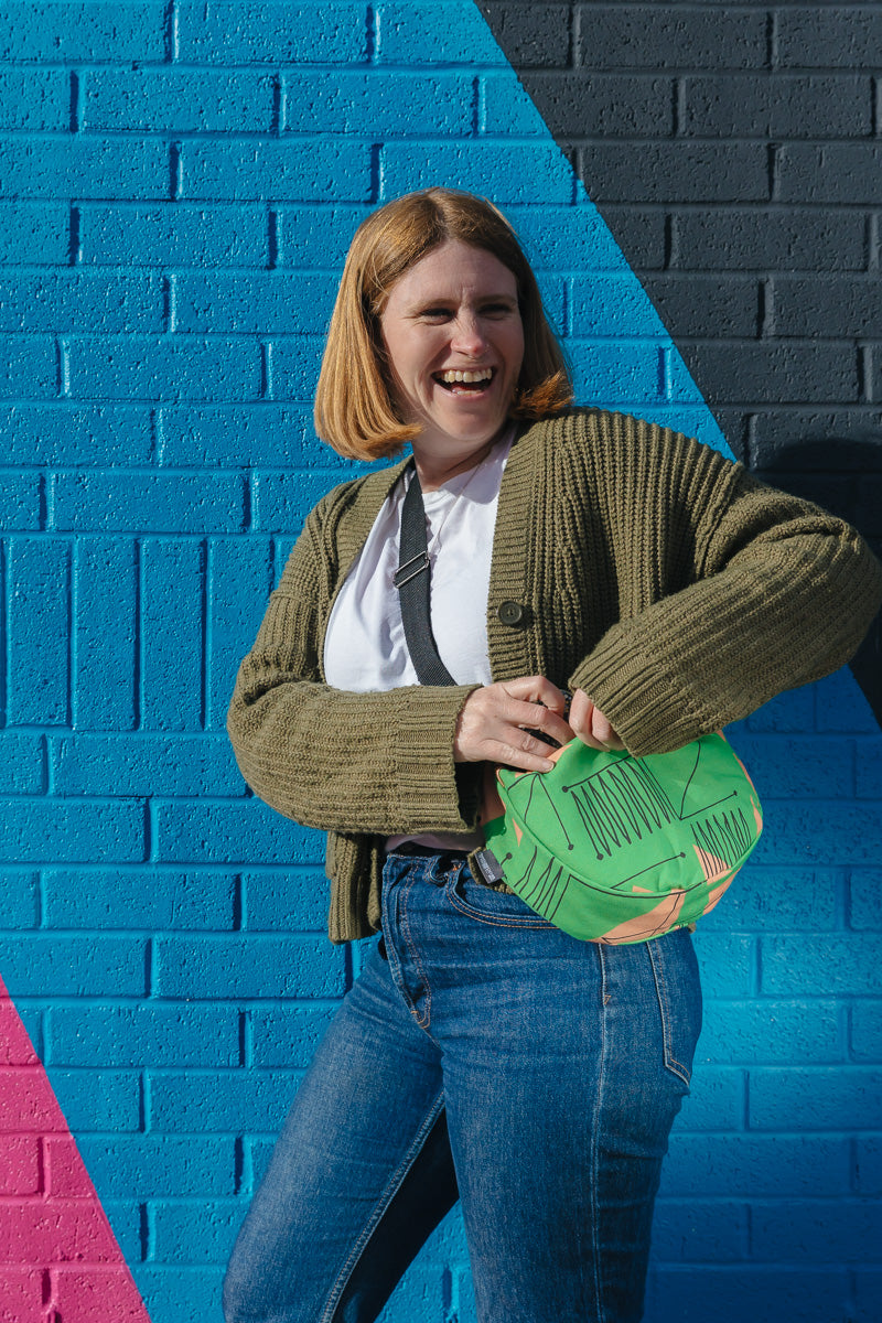 Person holding a green small cross body travel bag against a colorful geometric-patterned wall.