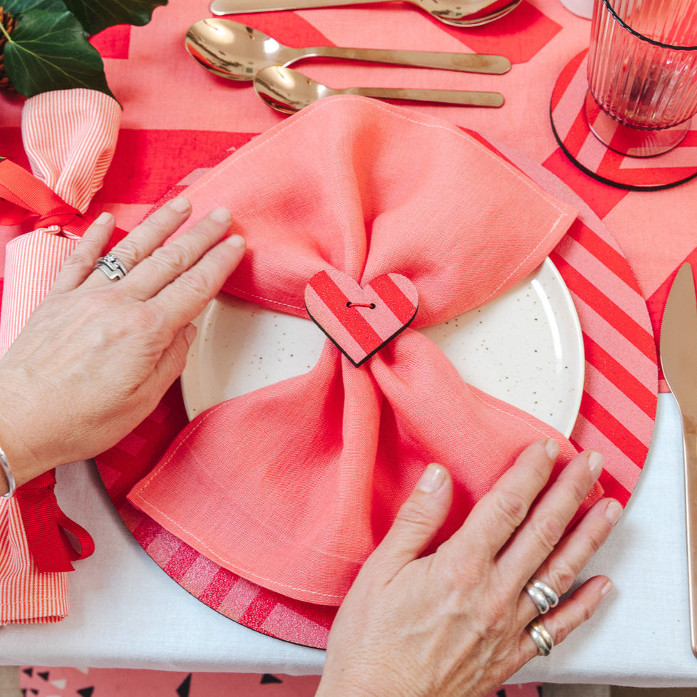 Person arranging a pink napkin on a table setting with cutlery and a festive background.