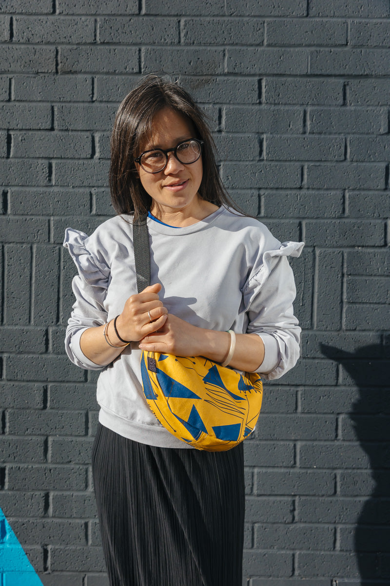Person wearing a white shirt with ruffled sleeves and a yellow bag against a gray brick wall.